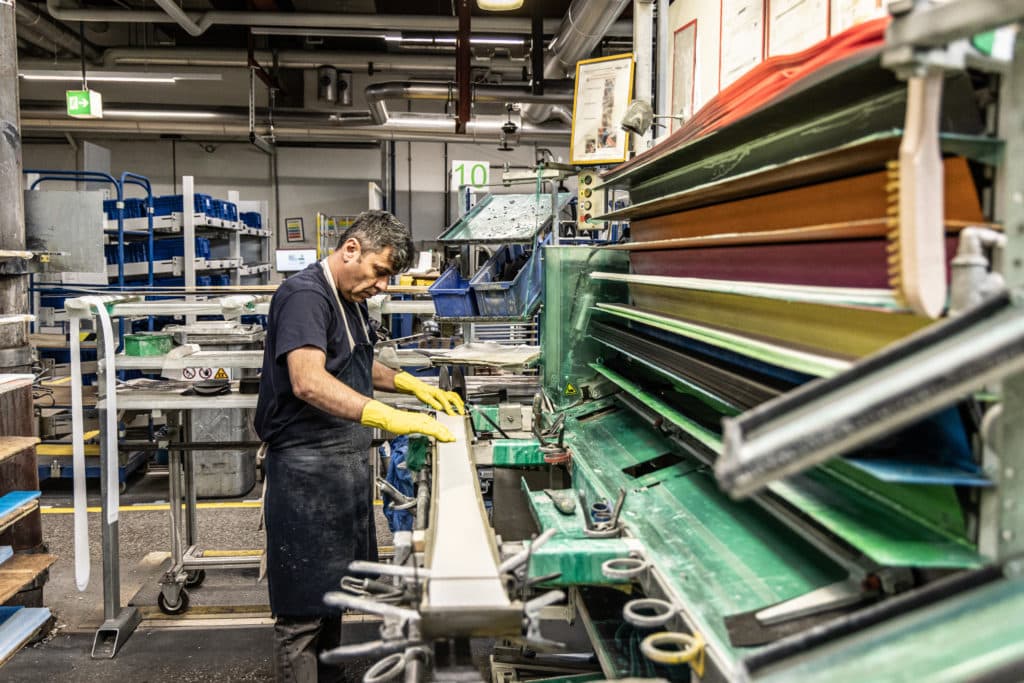 Ein Fabrikarbeiter mit gelben Handschuhen bearbeitet ein langes Werkstück an einer großen industriellen Fertigungsmaschine.