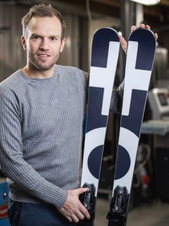 Ein Mann im grauen Pullover hält ein Paar Skier mit auffälligem schwarz-weißem geometrischem Muster.